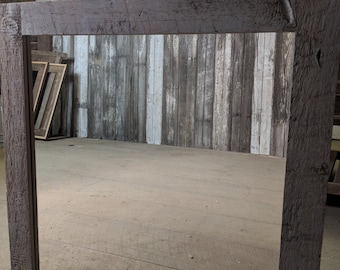 May include: A rustic wooden mirror frame with a lightly stained grey finish. The frame is made of reclaimed pine wood and has a rectangular shape. The mirror is reflecting a room with wooden walls.