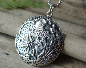 May include: A silver pendant necklace with a round, textured charm featuring a detailed design of stylized flowers and leaves.