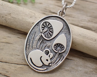 May include: A silver pendant featuring a detailed depiction of a mouse nestled amongst two mushrooms and blades of grass. The pendant is oval shaped and has a textured surface.