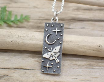 May include: A silver necklace with a rectangular pendant featuring a crescent moon, stars, and a moth. The pendant has a textured surface and is on a wooden background.