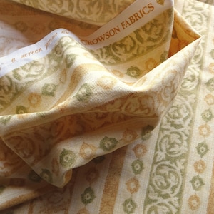 Könnte beinhalten: Cremefarbener Stoff mit einem sich wiederholenden Muster aus vertikalen Streifen und floralen Designs in Grün und Gold. Der Stoff ist mit "Crowson Fabrics" und "'Landerma' a screen print by" beschriftet.