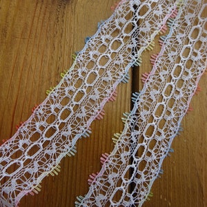 May include: Two strips of white lace with an openwork design, edged with small, rainbow-coloured accents. The lace is displayed on a wooden surface, highlighting its intricate pattern and colourful details. The lace is suitable for various craft projects.