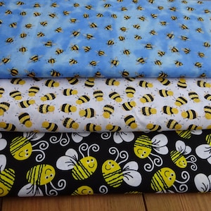 May include: Three fabric swatches with bee patterns. The top swatch is light blue with scattered yellow and black bees. The middle swatch is white with yellow and black bees. The bottom swatch is black with large yellow and white bees.