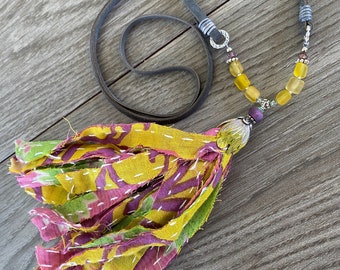 May include: A yellow, pink and green fabric tassel necklace with a brown leather cord and a silver chain with yellow glass beads. The necklace has a silver charm with a purple stone.