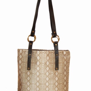 May include: A beige tote bag with a snakeskin pattern. The bag has dark brown leather handles attached with gold-colored rings. The handles are secured with small gold rivets.