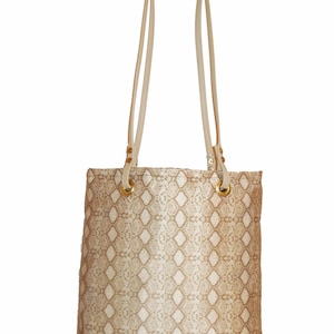 May include: A beige tote bag with a snakeskin pattern. The bag has long, cream-colored straps attached with gold-colored metal rings. The bag is rectangular with a flat bottom.