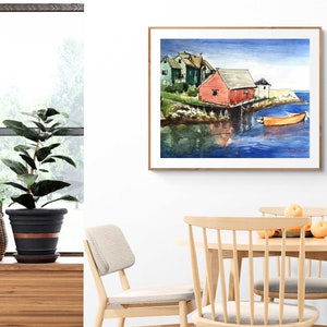 Puede incluir: Pintura acuarela de un paisaje mar&iacute;timo con una casa roja sobre pilotes y un peque&ntilde;o bote naranja en el agua. La pintura est&aacute; enmarcada en un marco de madera claro y est&aacute; colgada en una pared blanca.
