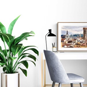 May include: A watercolor painting of a cityscape with a tall tower and a church in the background. The painting is framed in a brown wood frame and is hanging on a white wall above a white desk with a plant and a chair.