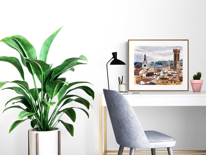 May include: A watercolor painting of a cityscape with a tall tower and a church in the background. The painting is framed in a brown wood frame and is hanging on a white wall above a white desk with a plant and a chair.