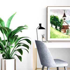 Puede incluir: Una pintura de acuarela de una peque&ntilde;a iglesia con un techo marr&oacute;n y un campanario blanco. La iglesia est&aacute; en una colina verde con &aacute;rboles en el fondo. La pintura est&aacute; enmarcada en un marco marr&oacute;n y est&aacute; colgada en una pared blanca.
