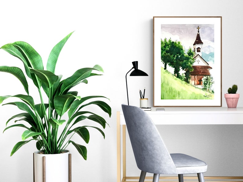 Puede incluir: Una pintura de acuarela de una peque&ntilde;a iglesia con un techo marr&oacute;n y un campanario blanco. La iglesia est&aacute; en una colina verde con &aacute;rboles en el fondo. La pintura est&aacute; enmarcada en un marco marr&oacute;n y est&aacute; colgada en una pared blanca.