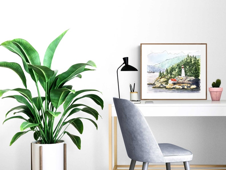 May include: A watercolor painting of a lighthouse on a rocky island with a red roof and white walls. The lighthouse is surrounded by green trees and a body of water. The painting is framed in a brown wood frame and is hanging on a white wall.