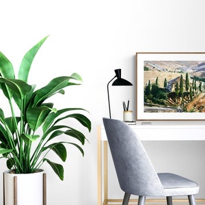 May include: A framed print of a watercolor painting depicting a landscape with a house and trees in a valley. The print is hanging on a white wall above a white desk with a plant, a lamp, and a chair.
