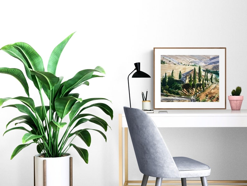 May include: A framed print of a watercolor painting depicting a landscape with a house and trees in a valley. The print is hanging on a white wall above a white desk with a plant, a lamp, and a chair.