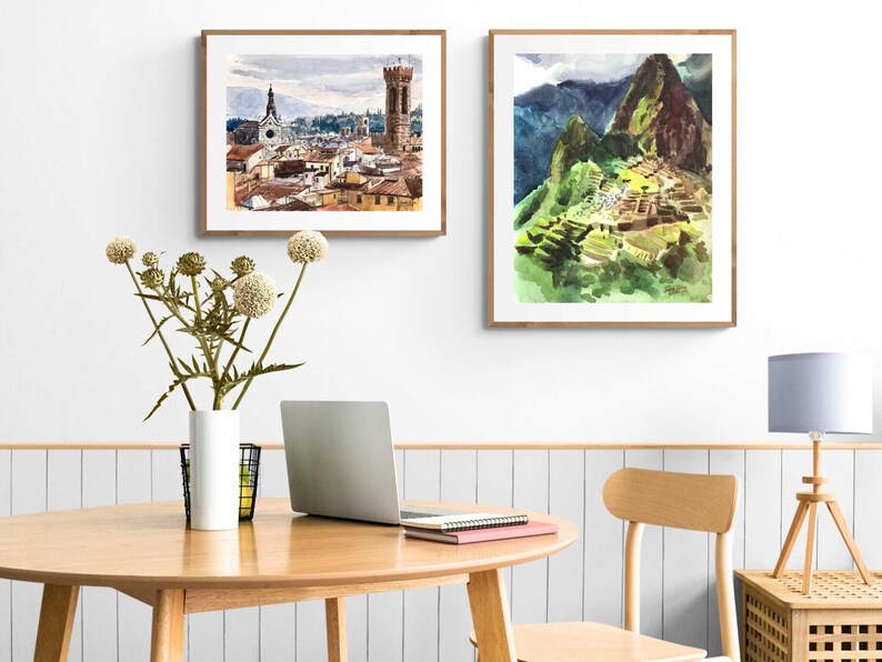May include: Two framed watercolor paintings, one depicting a cityscape with a tall tower and the other showing a mountain range with a stone structure.