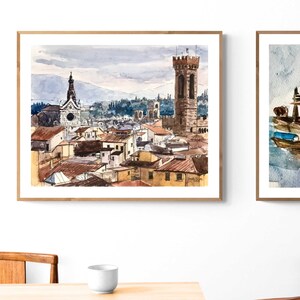 May include: A watercolor painting of a cityscape with a tall tower and a church in the background. The painting is framed in a light wood frame.