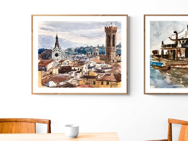 May include: A watercolor painting of a cityscape with a tall tower and a church in the background. The painting is framed in a light wood frame.