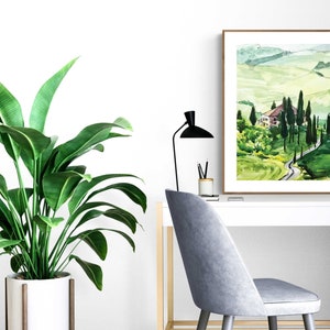 May include: A watercolor painting of a green landscape with a winding road leading to a small house. The painting is framed in a brown frame and is hanging on a white wall.