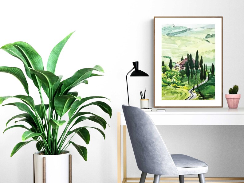May include: A watercolor painting of a green landscape with a winding road leading to a small house. The painting is framed in a brown frame and is hanging on a white wall.
