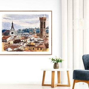 May include: A watercolor painting of a cityscape with a tall tower and a church in the background. The painting is framed in a light wood frame.