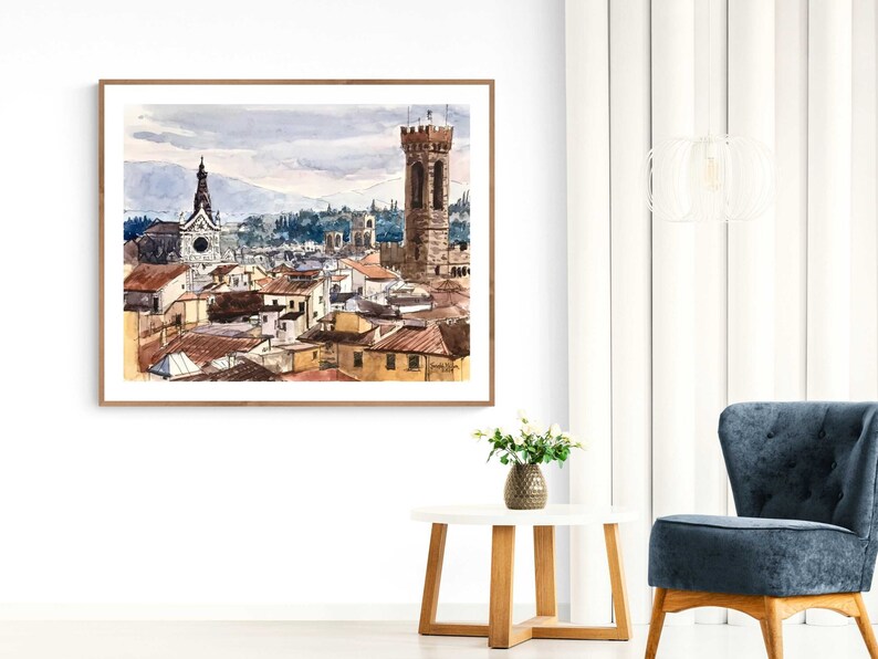 May include: A watercolor painting of a cityscape with a tall tower and a church in the background. The painting is framed in a light wood frame.