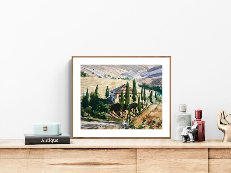 May include: A framed watercolor painting of a rural landscape with a house and trees. The painting is in a brown frame and is hanging on a white wall.