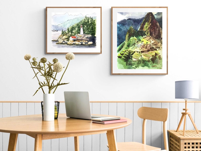 May include: Two framed watercolor paintings on a white wall. The painting on the left depicts a lighthouse on a rocky island with a forest in the background. The painting on the right depicts a mountain range with a stone structure built into the side of the mountain.