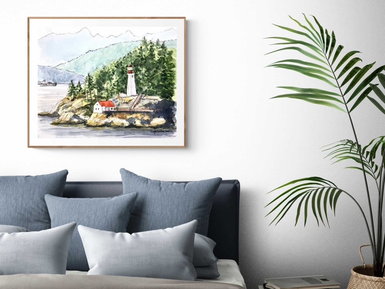 May include: Watercolor painting of a lighthouse on a rocky island with a red roofed building and green trees in the background. The painting is framed in a light wood frame.