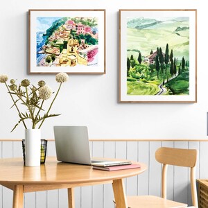 May include: Two framed watercolor paintings of Italian landscapes. The first painting depicts a colorful village on a cliff overlooking the sea. The second painting shows a winding road through rolling green hills.