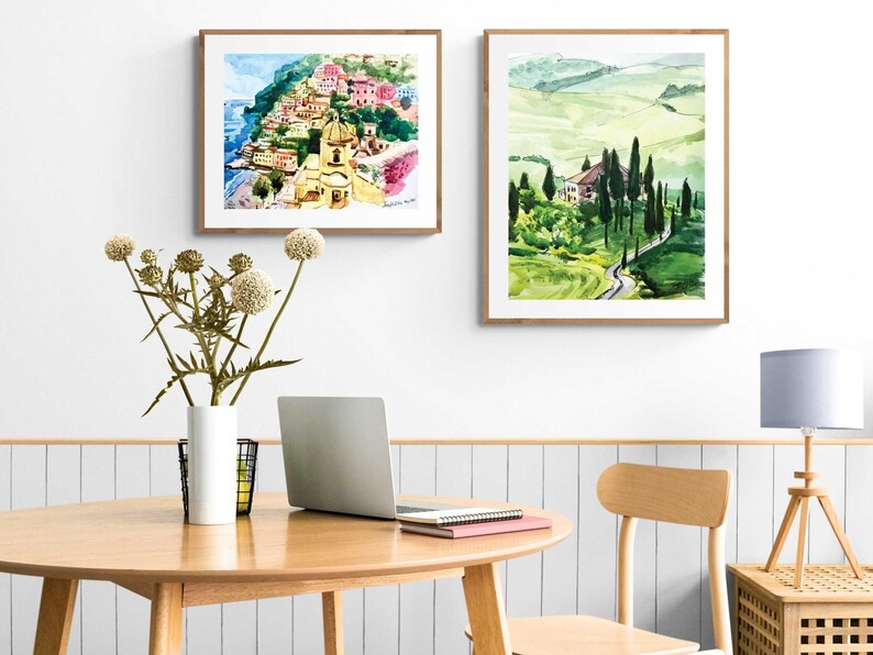 May include: Two framed watercolor paintings of Italian landscapes. The first painting depicts a colorful village on a cliff overlooking the sea. The second painting shows a winding road through rolling green hills.