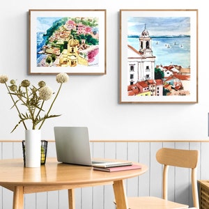Pu&ograve; includere: Due acquerelli incorniciati di paesaggi urbani europei. Il dipinto a sinistra mostra un paesaggio urbano colorato con una chiesa e una scogliera. Il dipinto a destra mostra una chiesa bianca con un cielo blu e acqua sullo sfondo.