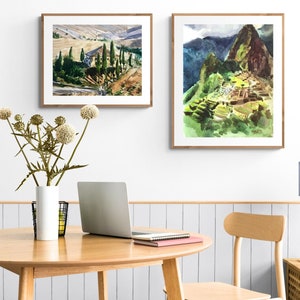 May include: Two framed watercolor paintings on a white wall. The painting on the left depicts a rural landscape with a building and trees. The painting on the right depicts a mountain range with a stone structure.