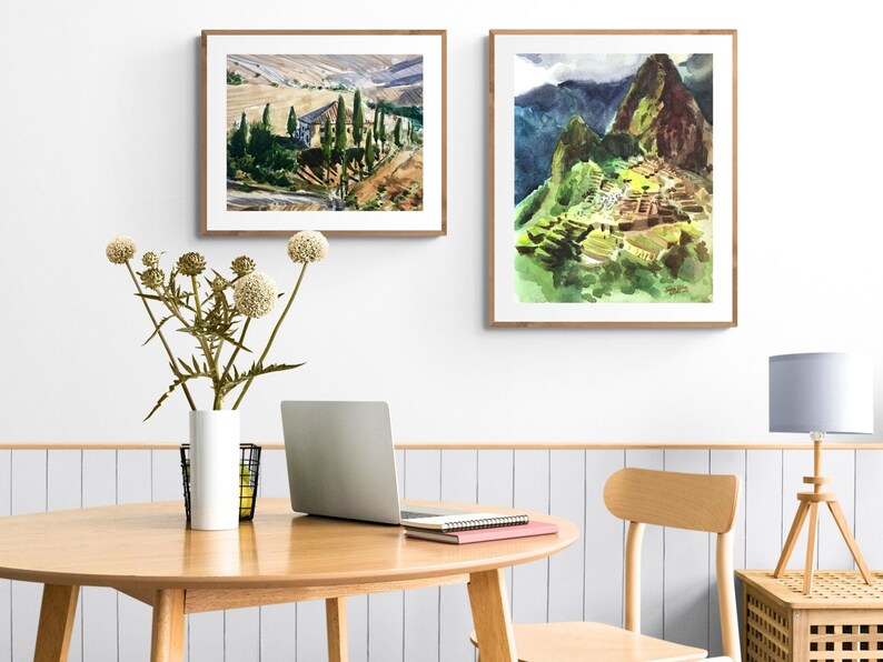 May include: Two framed watercolor paintings on a white wall. The painting on the left depicts a rural landscape with a building and trees. The painting on the right depicts a mountain range with a stone structure.