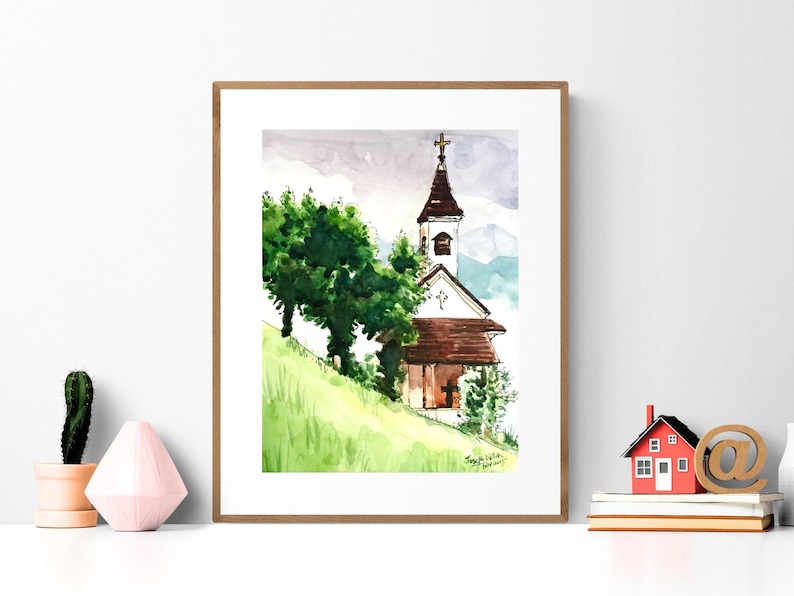 Puede incluir: Pintura acuarela de una iglesia blanca con un techo marr&oacute;n y una cruz en la parte superior. La iglesia est&aacute; rodeada de &aacute;rboles verdes y una colina cubierta de hierba. La pintura est&aacute; enmarcada en un marco de madera.
