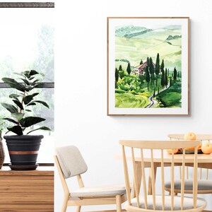May include: A watercolor painting of a green landscape with a winding road leading to a small house. The painting is framed in a light wood frame and is hanging on a white wall.