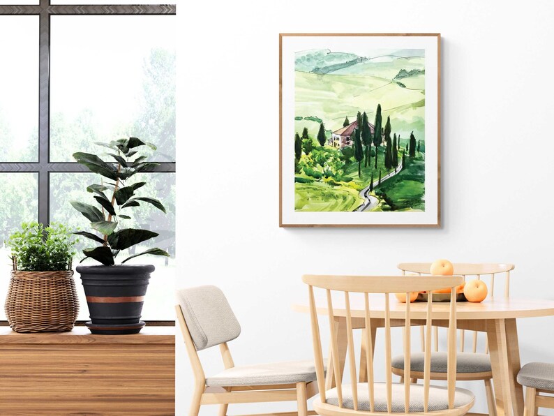 May include: A watercolor painting of a green landscape with a winding road leading to a small house. The painting is framed in a light wood frame and is hanging on a white wall.