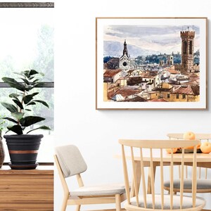 May include: A watercolor painting of a cityscape with a tall tower and a church in the background. The painting is framed in a light wood frame and is hanging on a white wall.