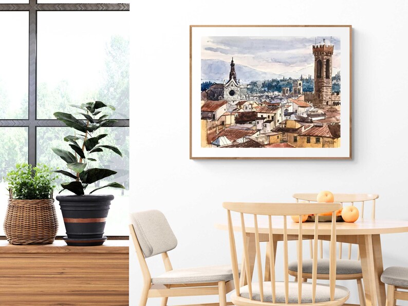 May include: A watercolor painting of a cityscape with a tall tower and a church in the background. The painting is framed in a light wood frame and is hanging on a white wall.