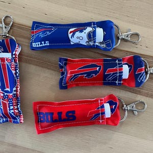 May include: Three different lip balm holders with keychains. Each holder is made of fabric with a Buffalo Bills theme. The first holder is blue and red with the team logo and the words "Bills" printed on it. The second holder is red and blue with a buffalo silhouette and the words "Bills" printed on it. The third holder is red with the words "Bills" printed on it.