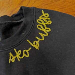 May include: Black crew neck sweatshirt with the words "Sko Bulls" embroidered in yellow thread across the upper chest. The embroidery has a cursive style. The sweatshirt is made of a soft material.