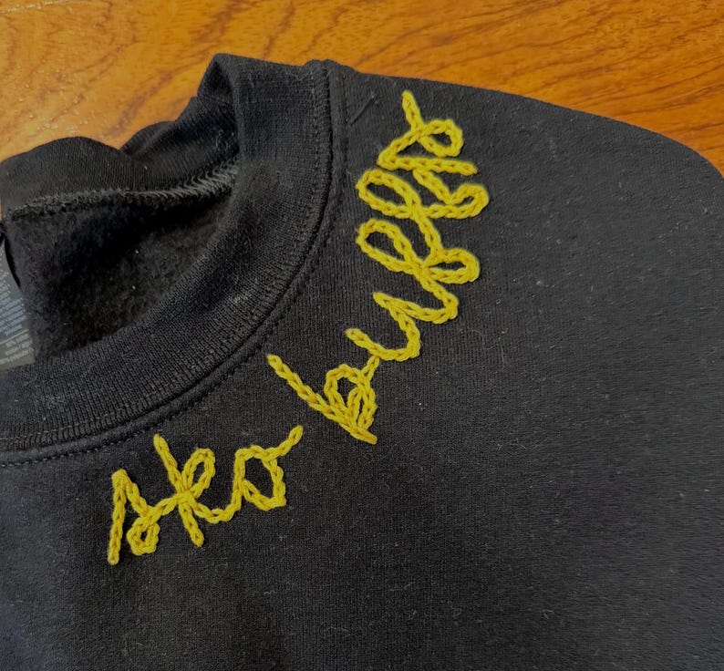 May include: Black crew neck sweatshirt with the words "Sko Bulls" embroidered in yellow thread across the upper chest. The embroidery has a cursive style. The sweatshirt is made of a soft material.