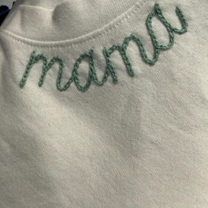 May include: White crew neck shirt with the word "mama" embroidered in green thread across the chest. The embroidery is in a cursive font. The shirt is made of a soft, light-colored fabric.