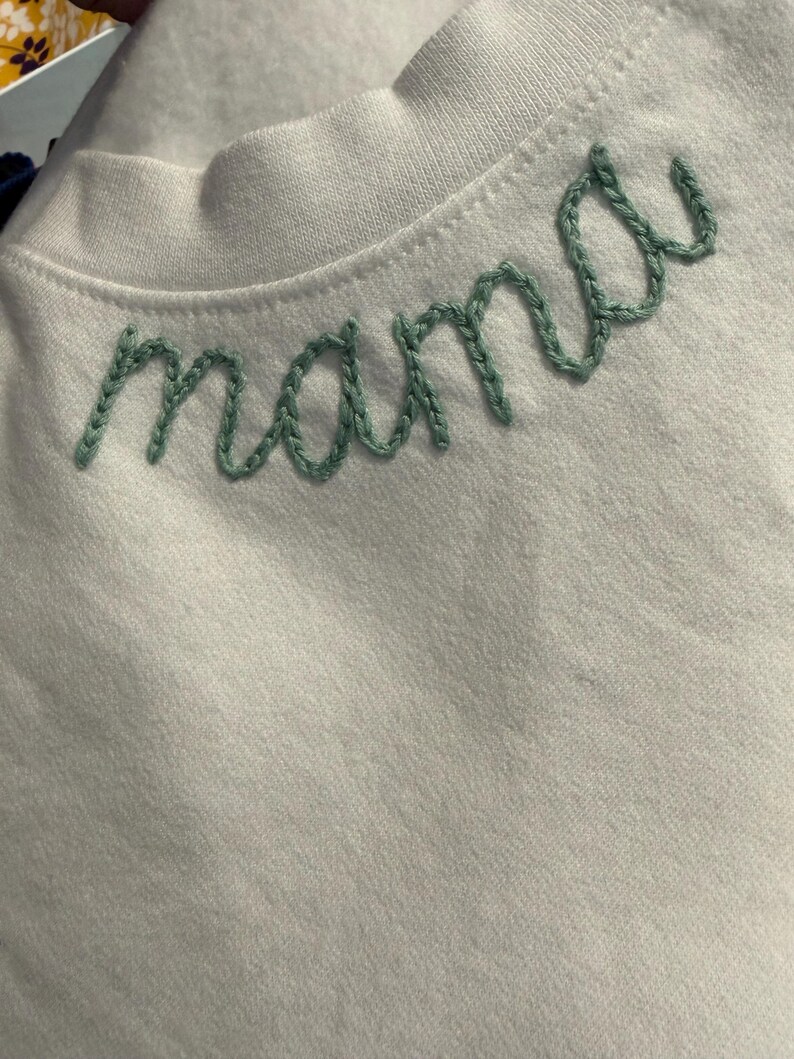 May include: White crew neck shirt with the word "mama" embroidered in green thread across the chest. The embroidery is in a cursive font. The shirt is made of a soft, light-colored fabric.