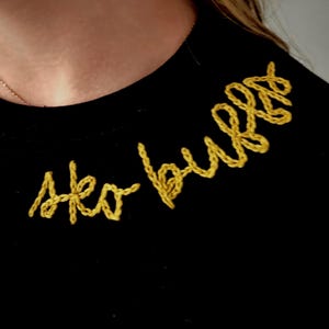 May include: Black crew neck shirt with the embroidered text "Ako bulls" in yellow thread. The shirt is made of a soft material and is suitable for casual wear.