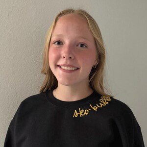 May include: A black sweatshirt with the embroidered text "sko bulla" in gold thread. The sweatshirt is worn by a person with blonde hair and a smiling expression. The background is a plain, light-colored wall.