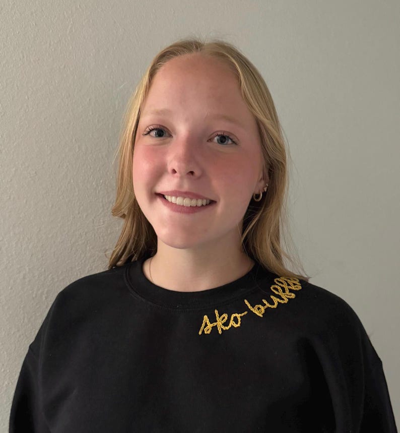 May include: A black sweatshirt with the embroidered text "sko bulla" in gold thread. The sweatshirt is worn by a person with blonde hair and a smiling expression. The background is a plain, light-colored wall.