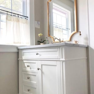 May include: White bathroom vanity with three drawers and a cabinet door. The vanity has brushed nickel hardware and sits on a black and white patterned tile floor. A gold framed mirror hangs above the vanity.