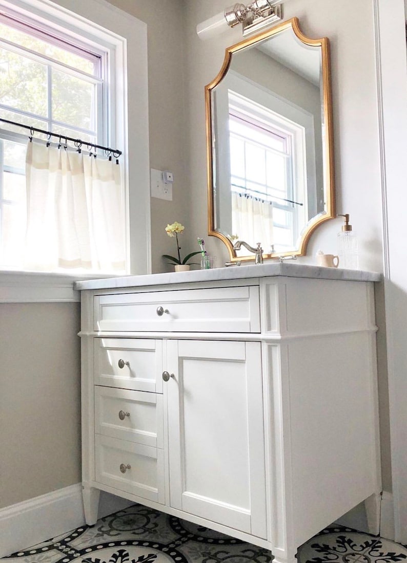 May include: White bathroom vanity with three drawers and a cabinet door. The vanity has brushed nickel hardware and sits on a black and white patterned tile floor. A gold framed mirror hangs above the vanity.
