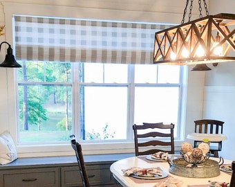 May include: A farmhouse style dining room with a window overlooking a lake. The window has a white and gray gingham roman shade. A wooden farmhouse style chandelier hangs above a table set with placemats and napkins.
