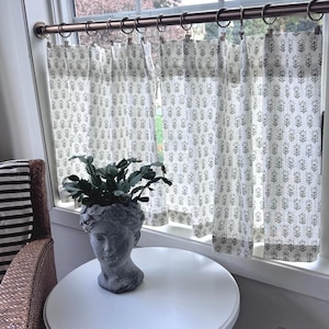 May include: A window with white curtains featuring a gray floral pattern. A gray stone head planter with a green plant sits on a white round table. The curtains are hung on a bronze rod with rings.